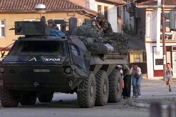 Einwohner von Prizren verstecken sich am 13.6.1999 bei einem Angriff serbischer Paramilitärs auf einen deutschen Kontrollpunkt hinter einem deutschen Panzerfahrzeug. Foto: picture-alliance / dpa | AFP Christophe Simon
