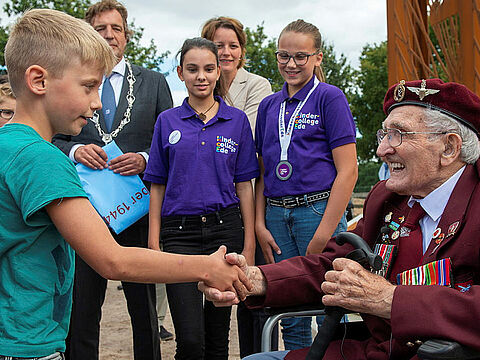 Kinder und Jugendliche bedanken sich beim 97-jährigen John Jeffries anlässlich der Einweihung einer Skulptur im Jahr 2019. Der Weltkriegsveteran und ehemalige Fallschirmspringer verstarb im Folgejahr. Foto: Ministry of Defense
