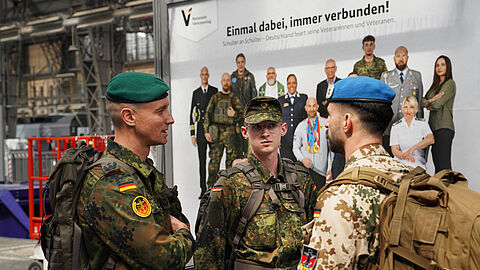 Oberstleutnant i.G. Marcel Bohnert (links) im Gespräch mit zwei reisenden Soldaten am Hauptbahnhof in Frankfurt am Main – im Hintergrund ein Plakat der Bundesregierung zum Nationalen Veteranentag am 15. Juni 2025. Foto: Lars Brinkmann/LCB Photography